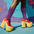 Coquette-style crew socks featuring a bold black background with hot pink and purple psychedelic squiggles, cherries, and shiny pink bows. Made of a soft polyester-spandex blend with reinforced black toe and heel. Fits US women's sizes 5–12 and men's up to 12. Stretchy, colorful, and cute for dopamine dressing.